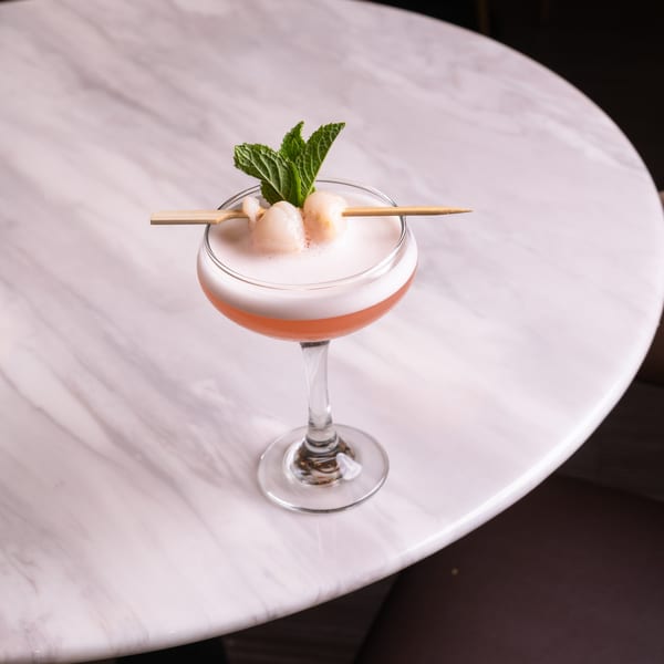 A foamy pink cocktail garnished with lychees and mint on a white marble table.