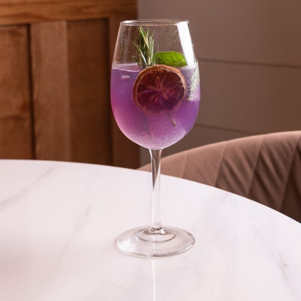 A vibrant purple cocktail with a dried orange slice, rosemary, and mint on a white marble table.