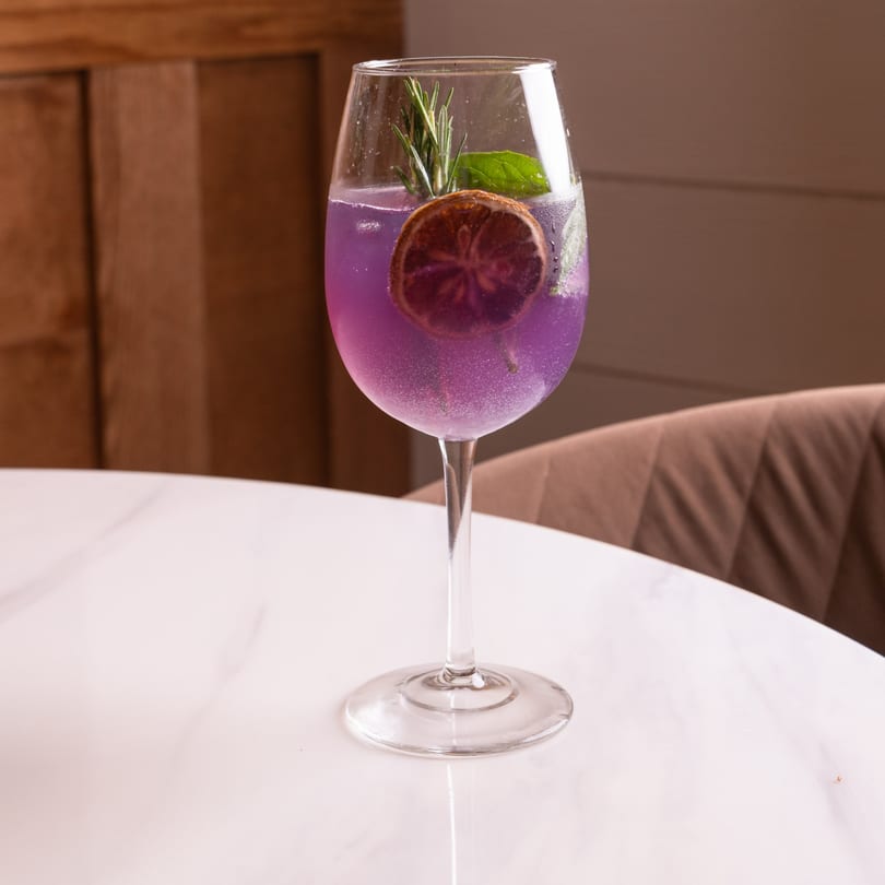 A vibrant purple cocktail with a dried orange slice, rosemary, and mint on a white marble table.