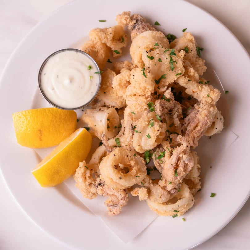 Crispy fried calamari with lemon wedges and a side of creamy dipping sauce on a white plate.