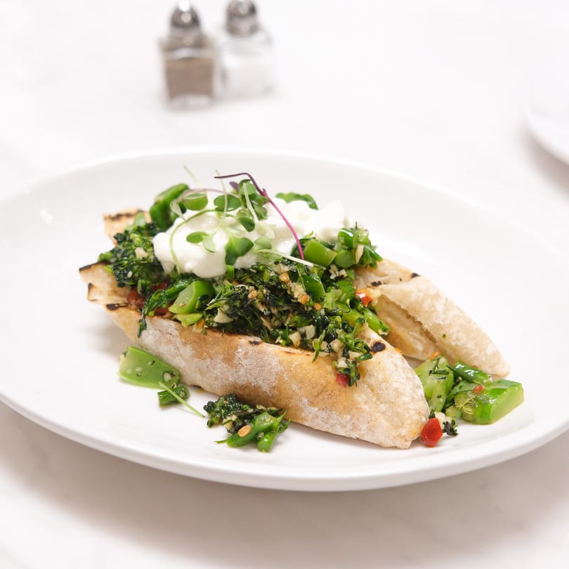 Italian bruschetta with green vegetables, white cheese, and microgreens on a white plate.