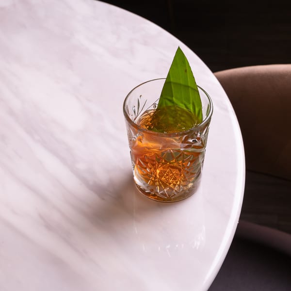 An amber cocktail in a cut glass with a green leaf garnish on a white marble table.