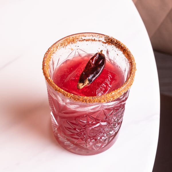 A red cocktail in a textured glass with a chili pepper garnish and a spiced rim.