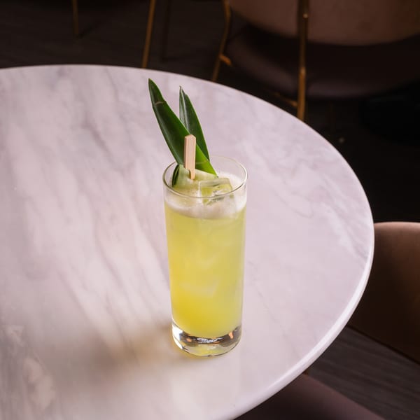 A tall glass of light green cocktail with ice and a green leaf garnish on a white marble table.